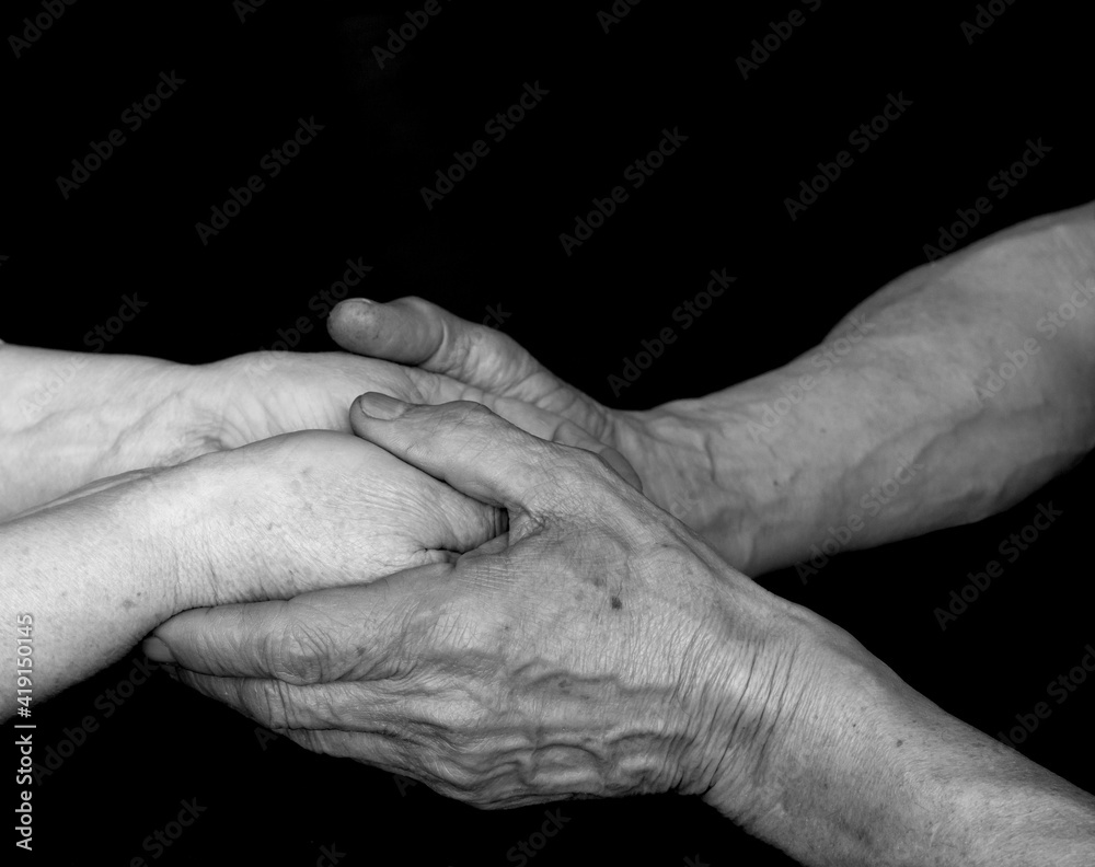 Fototapeta premium A black and white photo of an adult man and woman holding hands on a black background. Concept together forever. Elderly man and woman. Elderly couple holding hands