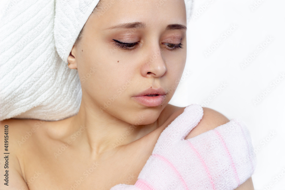 A young beautiful caucasian woman with a white towel on her head after a shower massaging her shoulder with an exfoliating hydro glove. Skin care, cosmetology