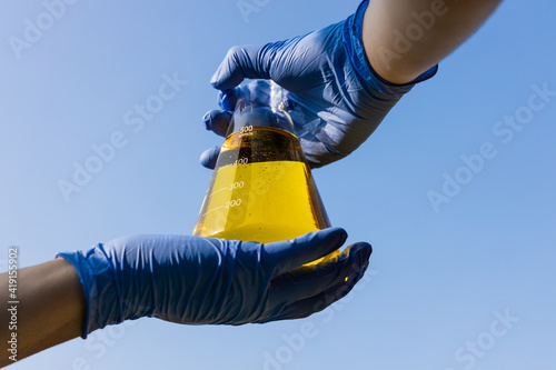 Hand with gloves holding beaker with ethanol biofuel against blue sky photo