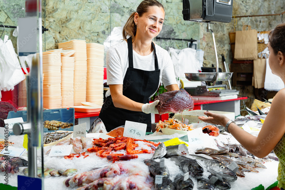 Polite glad cheerful positive female fishmonger standing behind counter of seafood store demonstrating raw bonito steak to woman buyer