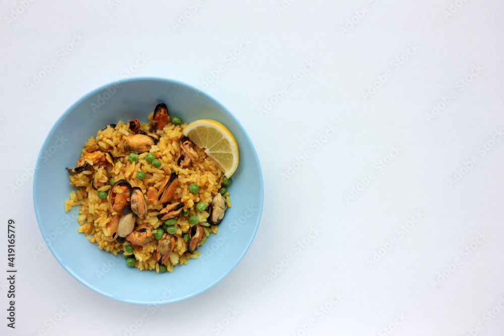 Traditional Spanish seafood dish paella with lemon slice on table. Mussel risotto with green peas on blue plate on white background. Top view, copy space. Healthy eating concept
