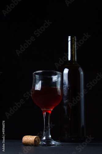 A glass of homemade wine next to a bottle on a dark background. Glass of red wine on a black background. Alcoholic drink