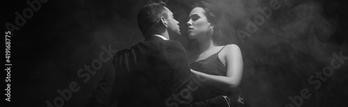 Monochrome shot of sensual woman looking at boyfriend in suit on black background with smoke, banner