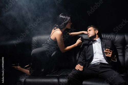 Man holding glass of whiskey near passionate woman in dress on couch on black background with smoke © LIGHTFIELD STUDIOS