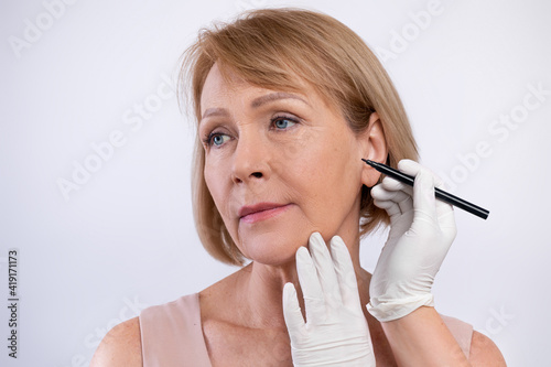 Doctor drawing marks on senior woman's face for cosmetic surgery operation on light studio background