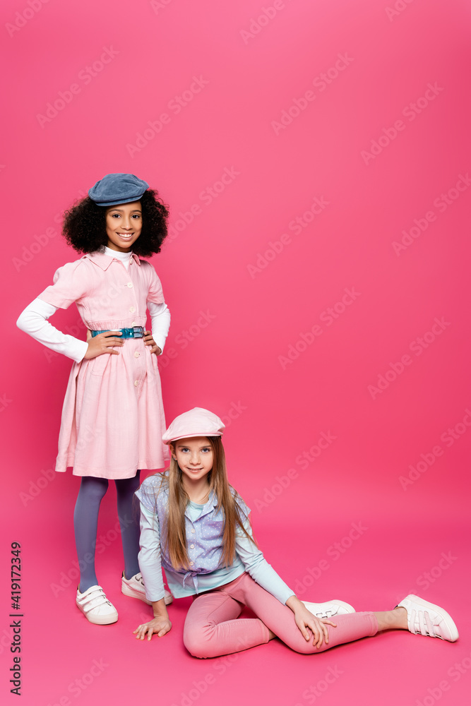 happy african american girl posing with hands on hips near friend sitting on pink