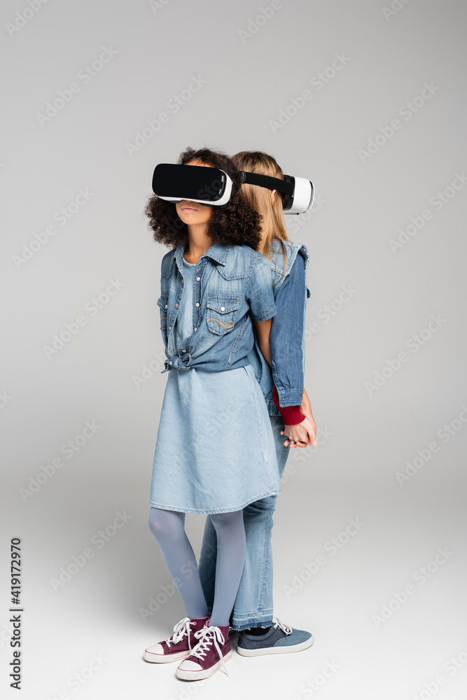 full length view of trendy interracial friends in vr headsets standing back to back on grey full length view of trendy interracial friends in vr headsets standing back to back on grey