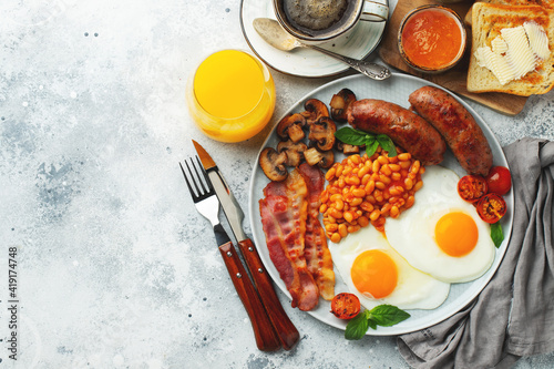 Full English breakfast on a plate with fried eggs, sausages, bacon, beans, toasts and coffee on light stone background. With copy space. Top view