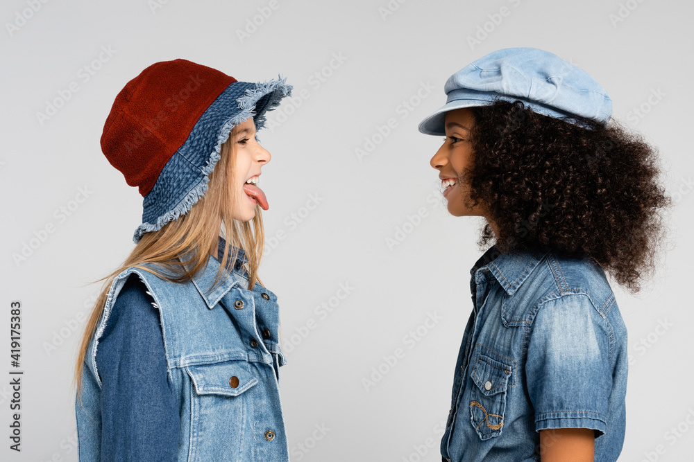 side view of cheerful girl sticking out tongue near happy african american friend isolated on grey side view of cheerful girl sticking out tongue near happy african american friend isolated on grey