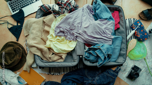 Top view on suitcase lay on floor with mess inside and around it. Clothes in pile on top of bag. Packing or unpacking in a rush. Hectic, stressed and anxious traveller