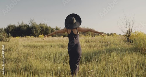 Beautiful mid adult woman in black hat walks through ukrainian steppe. She enjoys being at countryside under the sun. Woman wears black dress. Back view of undefined model, no face. A day in a life.