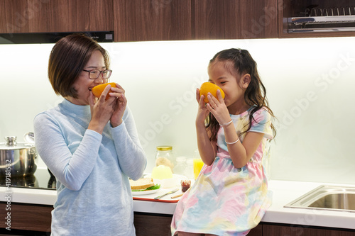 Happy Vietnamese mother and her little daughter eating delicious juicy oranges in kichen for breakfast