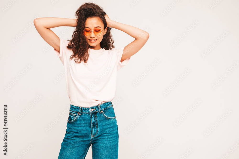 Beautiful black woman with afro curls hairstyle.Smiling model in white trendy jeans clothes. Sexy carefree female posing on grey background in studio in sunglasses. Tanned and cheerful Beautiful black woman with afro curls hairstyle.Smiling model in white trendy jeans clothes. Sexy carefree female posing on grey background in studio in sunglasses. Tanned and cheerful