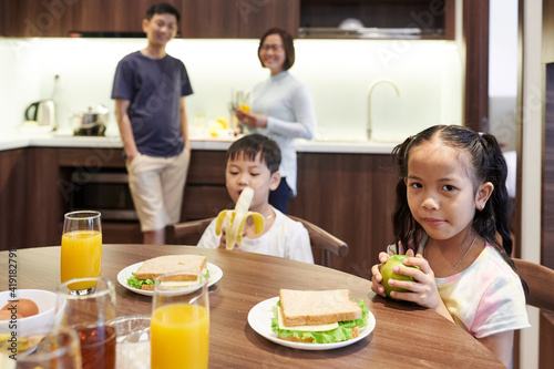 Preteen Vietnamese children sitting at kitchen table and eating sandwiches and fruits for breakfast
