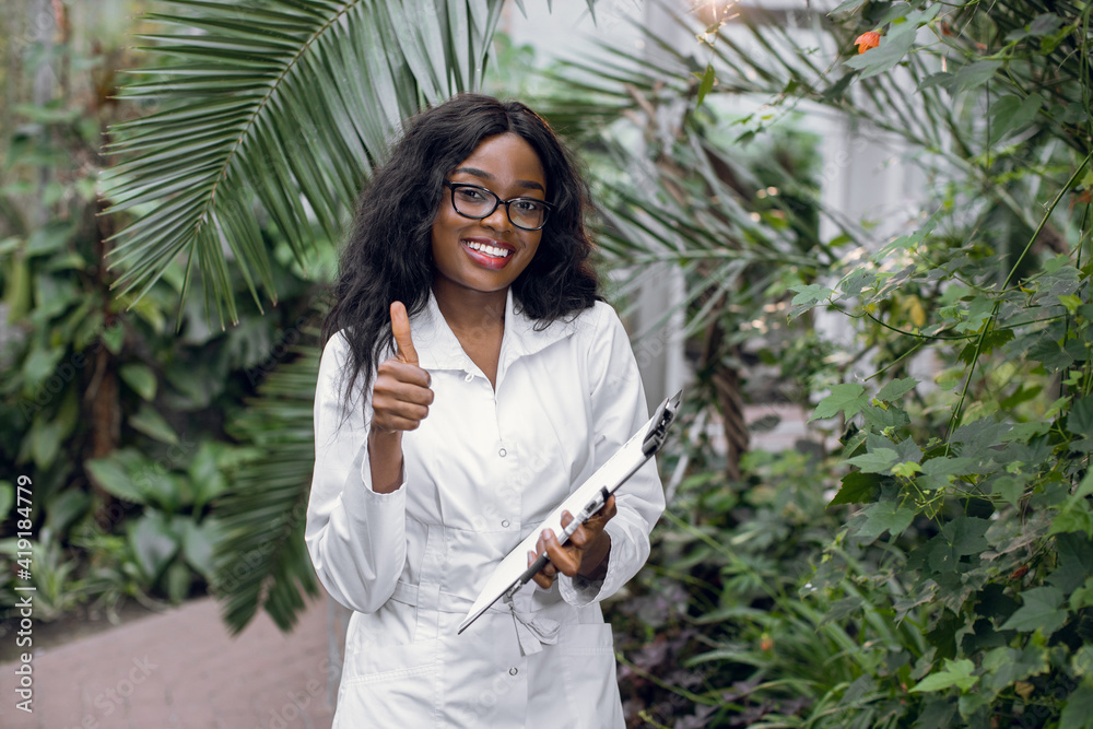 Young smiling African woman, professional scientist agronomist posing to camera with smile and thumb up, while standing in beautiful greenhouse with tropical plants. Young smiling African woman, professional scientist agronomist posing to camera with smile and thumb up, while standing in beautiful greenhouse with tropical plants.