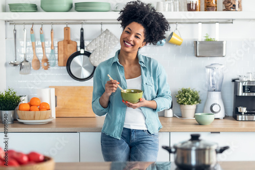 Beautiful afro woman eating noodles with chopsticks while looking at camera standing in the kitchen at home.