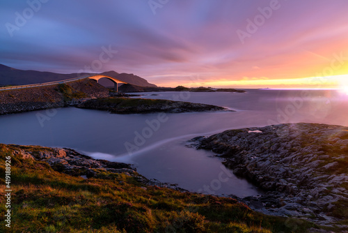 Vibrant sunset illuminates the arch of the famous great atlantic highway in northern norway. Long exposure with powerful light flares. Purple and orange sunset colors.