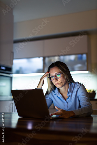 Pretty, middle-aged woman working late in the day on a laptop computer at home, running a business from home, working remotely - getting frustrated