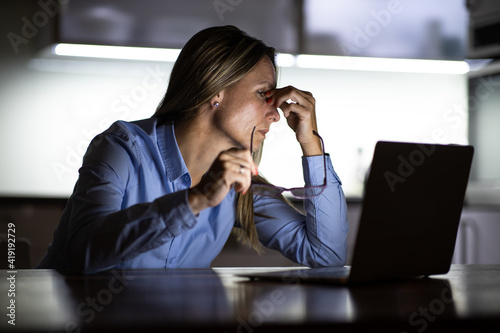 Pretty, middle-aged woman working late in the day on a laptop computer at home, running a business from home, working remotely - getting frustrated, exhausted