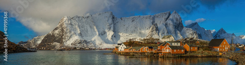 View of the picturesque fishing village of Reine with orange and red rorbu in the foreground and snow-capped mountain peaks in the background, Lofoten, Norway.