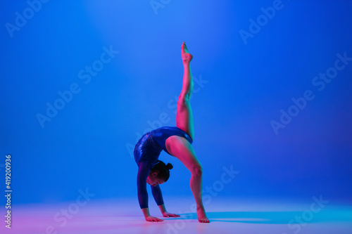 Agility. Young flexible girl isolated on blue studio background in neon light. Young female model practicing artistic gymnastics. Exercises for flexibility, balance. Grace in motion, sport, action.