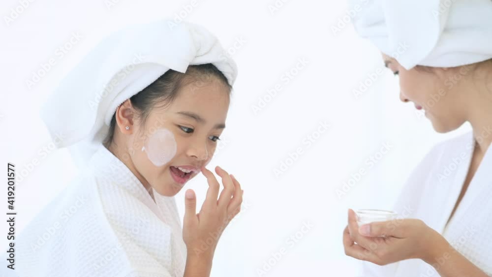 Happy beautiful mom and daughter in white bathrobe applying moisturizing cream on face at home, skin care and treatment concept..