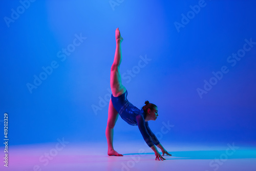 Agility. Young flexible girl isolated on blue studio background in neon light. Young female model practicing artistic gymnastics. Exercises for flexibility, balance. Grace in motion, sport, action.