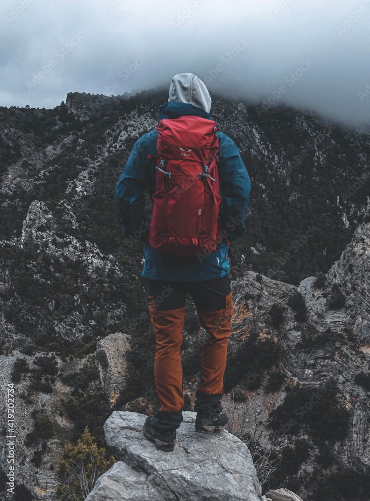 Boy who is traveling between the mountains, is traveling a path and it is dark, with bad weather, mysterious landscape. He is wearing red bag and blue jacket.