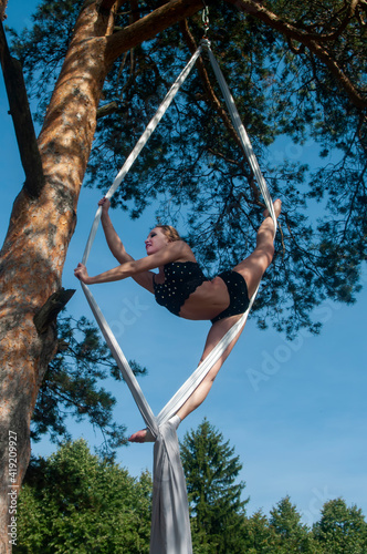 A slender girl with blonde hair, in a black stage costume, performs gymnastic and circus exercises on white silk against the background of the forest and the sky.