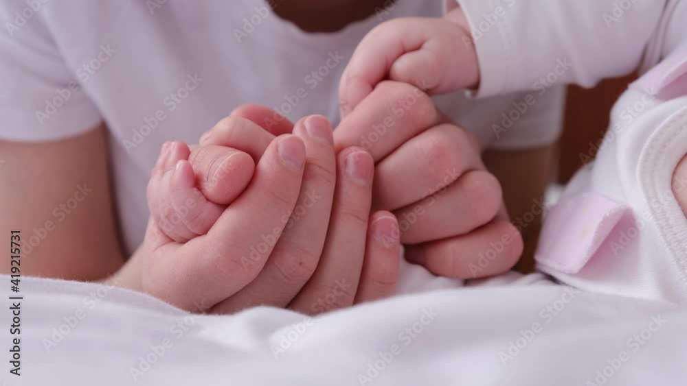 Close up of loving young mother gently hugs newborn baby daughter, holds and kisses hands lying together on cozy white bed at home, enjoying good moments. Gentle parenting and family love concept Close up of loving young mother gently hugs newborn baby daughter, holds and kisses hands lying together on cozy white bed at home, enjoying good moments. Gentle parenting and family love concept