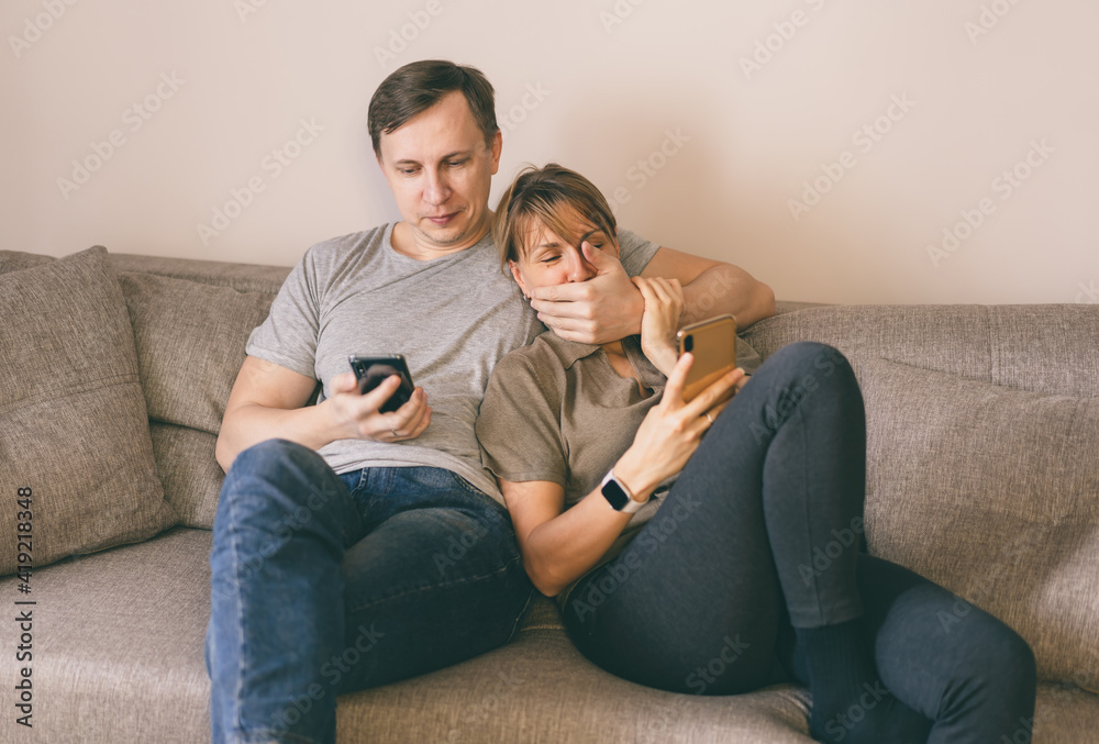 Man and woman sitting on a couch and holding smartphones in their hands. Man trying to close his wife mouth as a joke