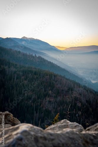 Vertical view of the setting sun in Tatra National Park, Poland. Coniferous forest, rocky peaks and orange light illuminating the sky. Selective focus on the summit, blurred background.