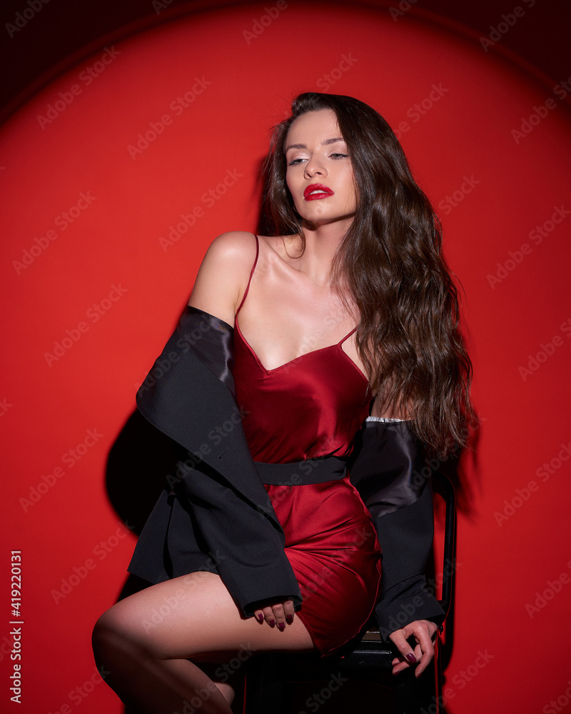 Young beautiful sexy woman with make-up and wavy brunette hair in red silk  dress posing against red background in circle of light (spot gobo light)  Stock-Foto | Adobe Stock