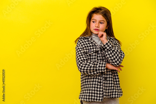 Little caucasian girl isolated on yellow background looking sideways with doubtful and skeptical expression.