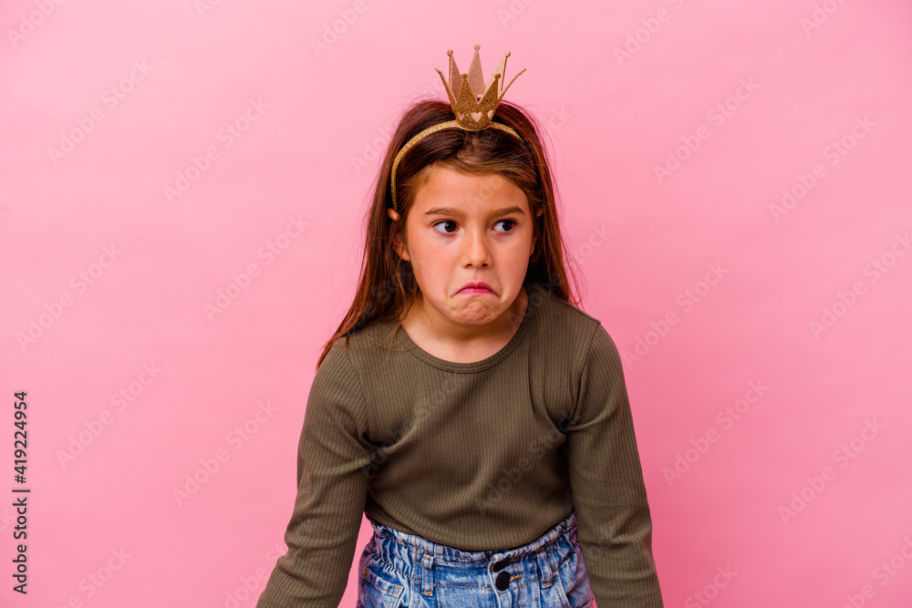 Little princess girl with crown isolated on pink background shrugs shoulders and open eyes confused.