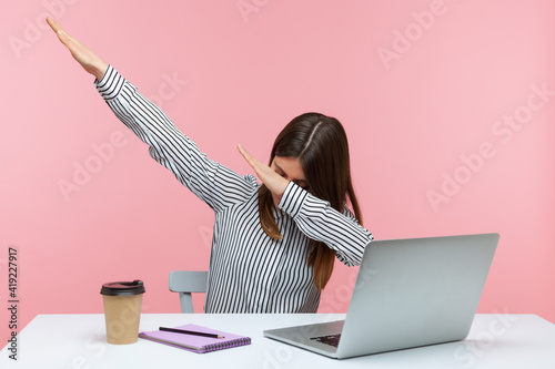 Extremely excited happy woman office worker showing dab dance gesture, performing internet meme of success, sitting at workplace with laptop. Indoor studio shot isolated on pink background photo
