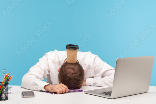 Fototapeta Overworked exhausted man office worker lying on table, paper coffee cup standing on his head, lack of energy, procrastination and professional burnout