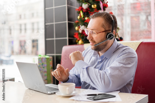 A middle-aged caucasian man with a headset in eyewear talking via video link with smile. He works during a coffee break with a laptop, mobile phone and papers on the table sitting in a cafe. SDOF photo