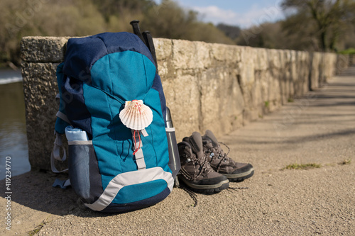 Backpack with seashell symbol of Camino de Santiago, trekking boots and poles leaning on stone wall. Pilgrimage to Santiago de Compostela. Copy space photo