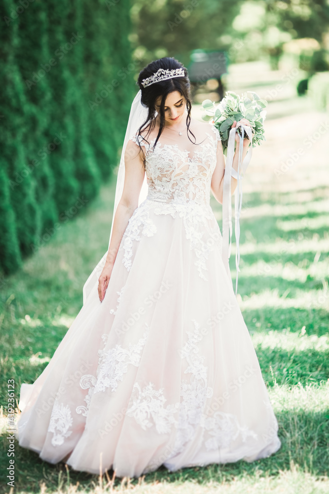 Beautiful brunette bride in elegant white dress holding bouquet posing neat trees