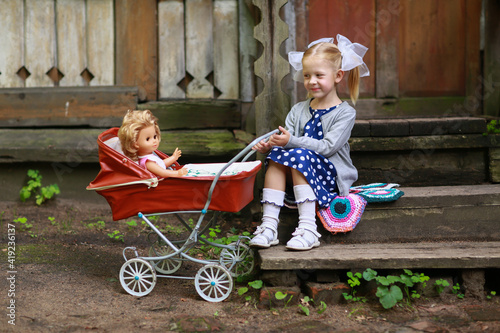 Cute smiling girl with bows in a blue dress playing with her red toy stroller and doll sitting on the porch of the house . Childhood in the Soviet Union