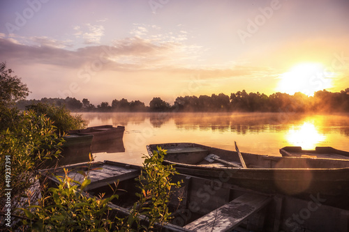 Beautiful countryside morning foggy scenery with boats on river with tranquil water and reflection during sunrise in warm colors during golden hour