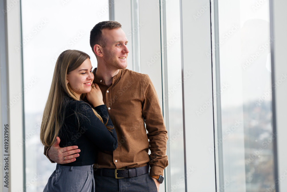 Stylish young couple in love in front of large panoramic windows. Young woman and man stand near a large window Stylish young couple in love in front of large panoramic windows. Young woman and man stand near a large window