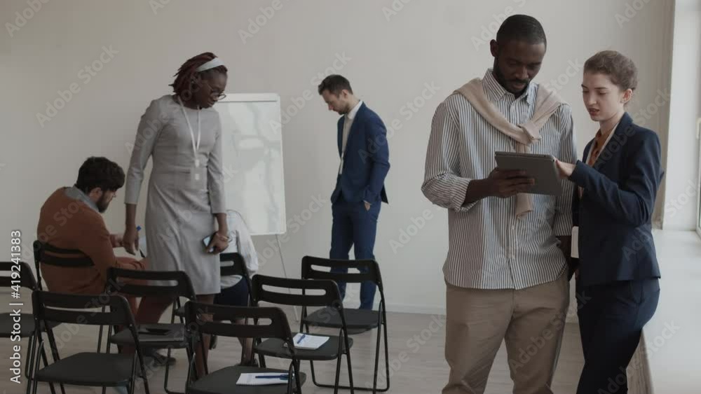 Medium long of African man and Caucasian woman dressed formally standing in conference room, in foreground of busy colleagues, using tablet computer and talking Medium long of African man and Caucasian woman dressed formally standing in conference room, in foreground of busy colleagues, using tablet computer and talking