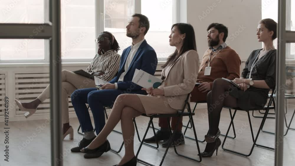Full side view of multiethnic female and male professional people dressed formally, sitting on chairs in large room with windows, looking at and listening to invisible conference speaker Full side view of multiethnic female and male professional people dressed formally, sitting on chairs in large room with windows, looking at and listening to invisible conference speaker