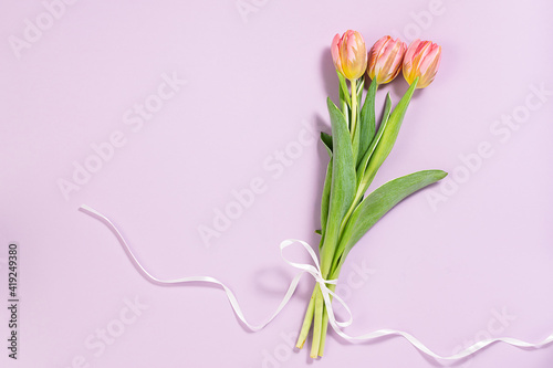 Fototapeta Naklejka Na Ścianę i Meble -  Minimalism style composition with beautiful pink tulips with ribbon on lilac pastel background top view. Spring greeting card template.