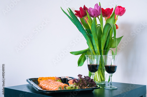 Bouquet of tulips in glass vase. Delicious sushe and rolls set. Japanese cuisine. Couple glasses with wine. Black table. White wall.