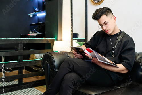 boy dressed in black sits drinking coffee while waiting for his turn in a barber shop © Luisphtos