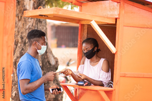 nigerian woman in a pos service kiosk attending to a man who wants to withdraw money, both wearing face mask
