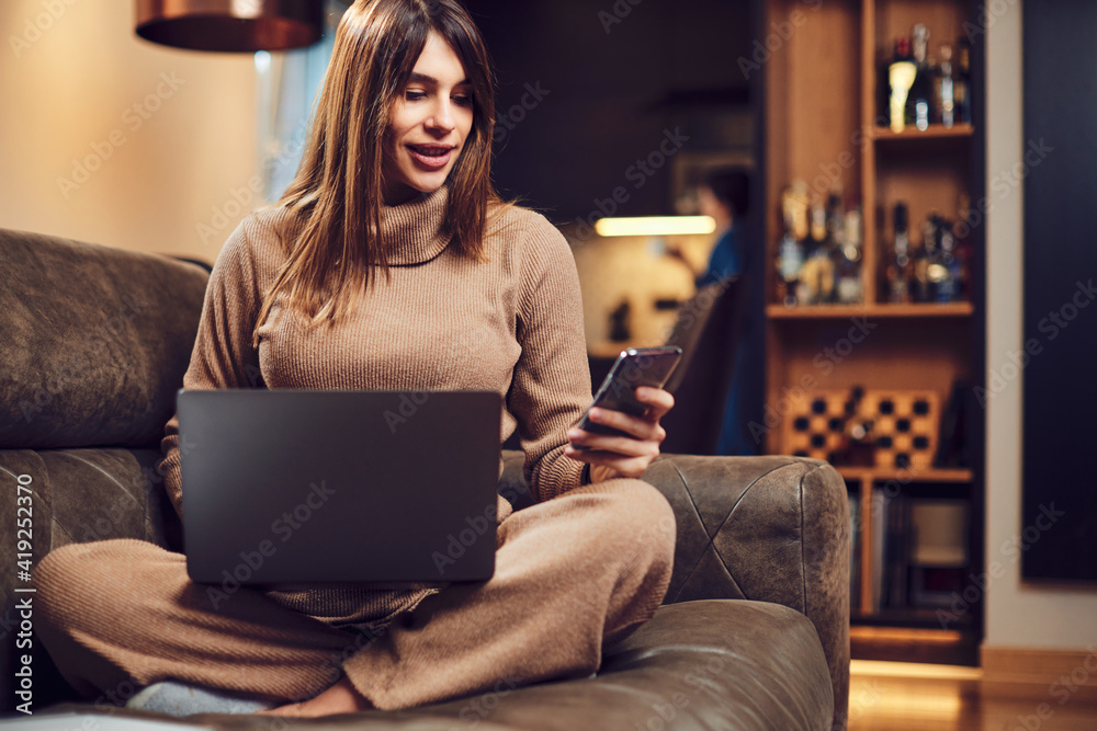 A pregnant brunette sitting on couch at home with laptop in lap and phone in hands and ordering something online. A pregnant brunette sitting on couch at home with laptop in lap and phone in hands and ordering something online.